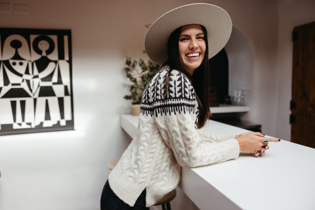 Jennifer Berg, a Navajo Native American woman, stands in her home smiling while wearing a beautifully knitted sweater.