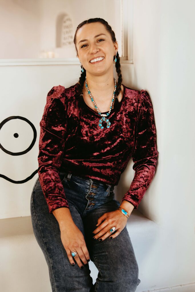Jessica Jonkman, a Navajo Native American woman, sits down and smiles while she wears a variety of beautifully crafted native american jewlery made with eye-catching turquoise.